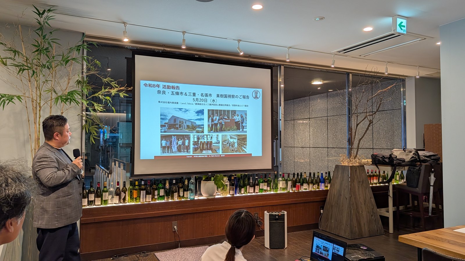 ワインボトルが並ぶ店内で、果樹園視察の報告スライドを前に話す農産物加工協会の発表者
